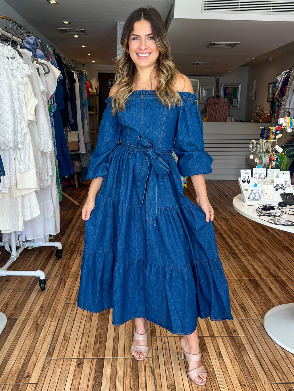 Medium indigo washed light weight denim midi dress featuring off shoulder neckline with ruffled edge, long sleeve, self sash tie, tiered skirt and smocked back bodice.