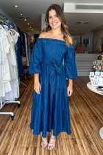 Load image into Gallery viewer, Medium indigo washed light weight denim midi dress featuring off shoulder neckline with ruffled edge, long sleeve, self sash tie, tiered skirt and smocked back bodice.
