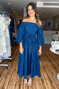 Medium indigo washed light weight denim midi dress featuring off shoulder neckline with ruffled edge, long sleeve, self sash tie, tiered skirt and smocked back bodice.